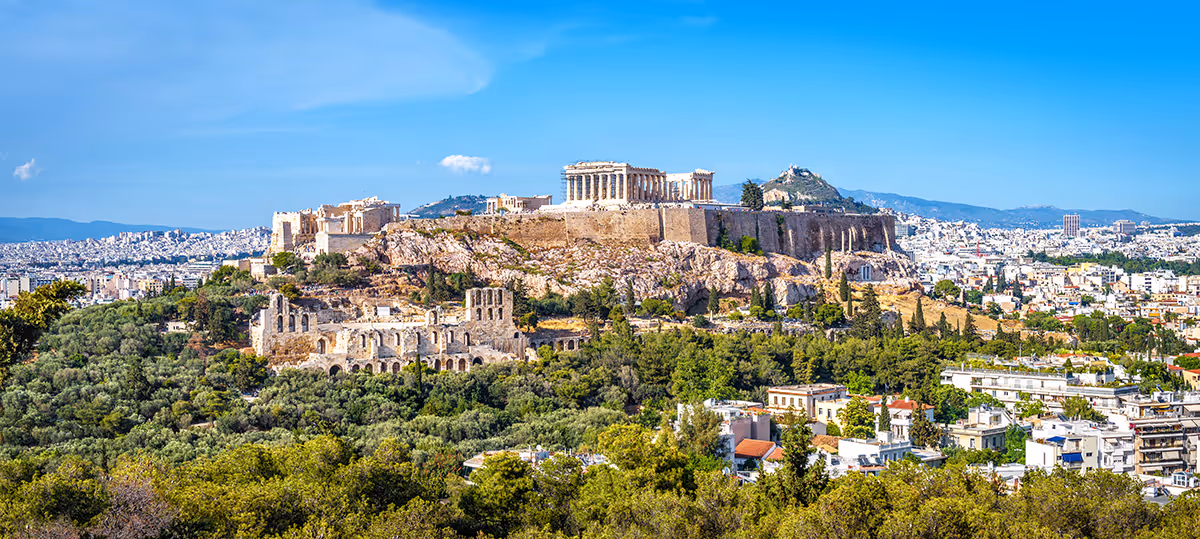 Blick auf die Akropolis von Athen mit dem Parthenon-Tempel auf einem felsigen Hügel über der Stadt und grün bewachsenen Flächen im Vordergrund.