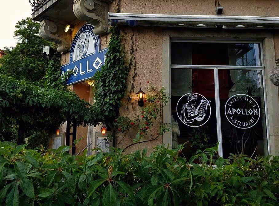 Außenansicht des griechischen Restaurants Apollon mit blauem Schild, Fenster mit Logo und grüner Bepflanzung.